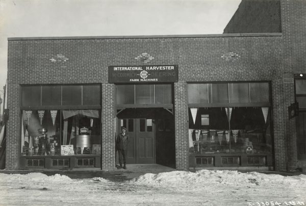 A man, possibly proprietor Charles C. Matezcvk(sp?), stands in the doorway of an International Harvester dealership. Advertising pennants and farm machines are visible through the front windows.