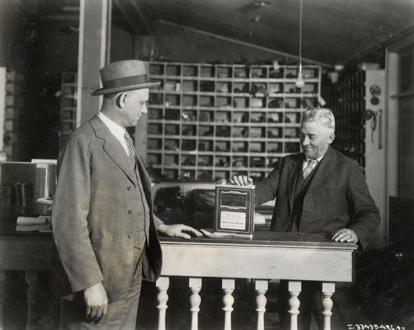 A man, possibly an International Harvester salesman, presents a can of cream separator oil to a customer. The setting is likely an International Harvester dealership. The text on the oil can reads: "Insist on This Oil."