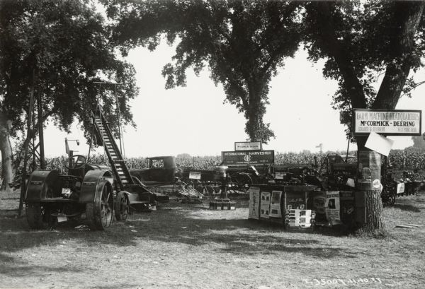 Signs nailed to trees advertise an assortment of agricultural equipment set up on outdoor display at the Schairer and Feyerherm farm machinery headquarters. Included in the display are a McCormick-Deering 10-20 tractor and numerous advertising posters. Schairer and Feyerherm were International Harvester dealers.