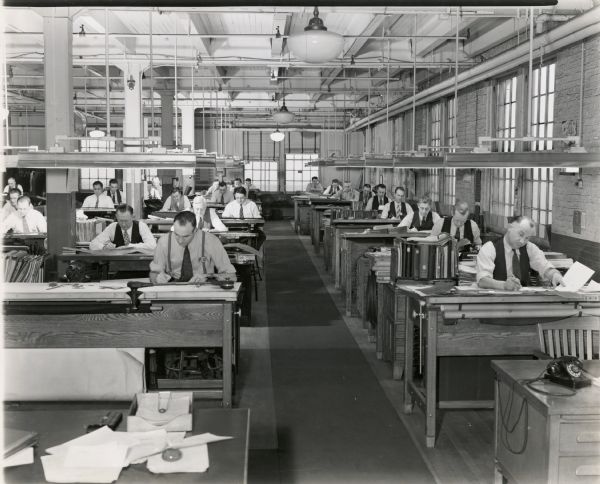 Men sit behind desks in the drafting room of International Harvester Company's farm machinery engineering department. The original caption reads: "A portion of the drafting room of International Harvester Company's farm machinery engineering department at Chicago. Here the designers put on paper the ideas which eventually are incorporated through the medium of steel and other materials into Harvester products."