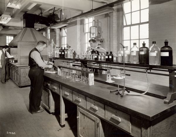 Men stand at the counter of a physical and chemical laboratory. The original caption reads: "A complete modern physical and chemical laboratory, a portion of which is shown in this photo, is operated in connection with one of the company's plants building an important war product. This laboratory is used for the testing of the materials used in the manufacture of the war product."