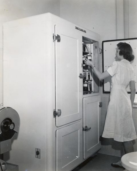 A woman stands next to a "Tomac Plasma Bank," a refrigerated cabinet designed to store blood plasma. The original caption reads: "Tomac Plasma Bank, engineered by International Harvester engineers in cooperation with the Baxter Laboratories of the American Hospital Supply Corporation near Chicago.  It is the only plasma bank with the convenient upright cabinet."
