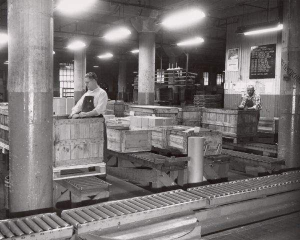 Two men are looking through wooden shipping crates in a room at International Harvester's Tractor Works.