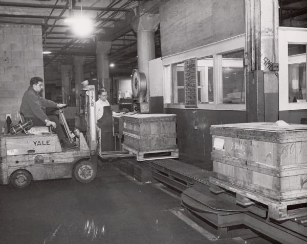 A man is using a forklift to load a wooden crate onto a conveyor belt at International Harvester's Tractor Works.