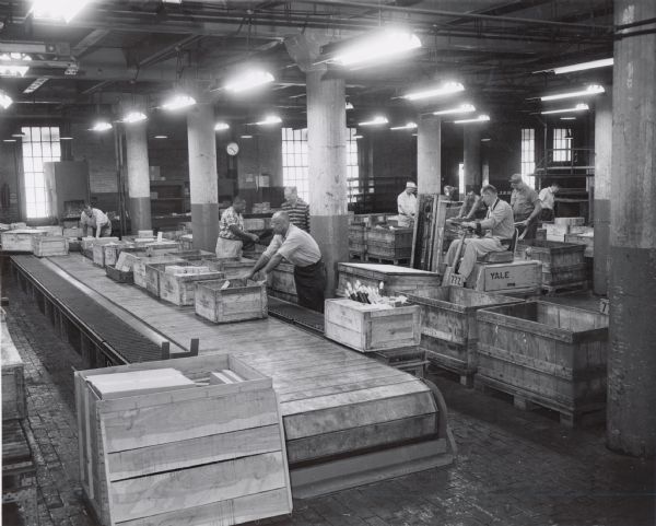 Two men are moving boxes along a conveyor belt at International Harvester's Tractor Works, while others are working in the background.