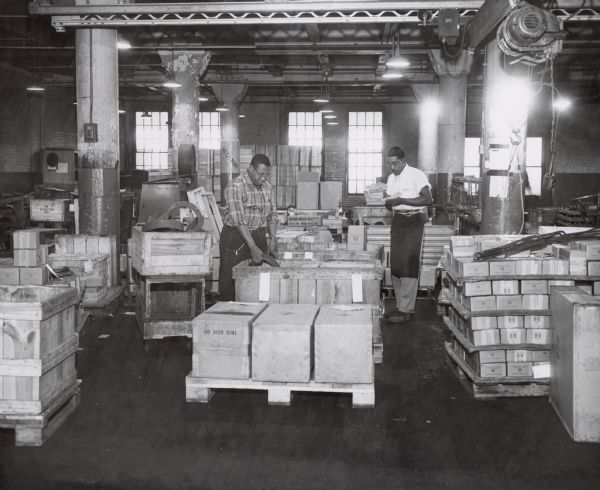 Two factory workers are loading (or unloading) wooden shipping crates at International Harvester's Tractor Works.