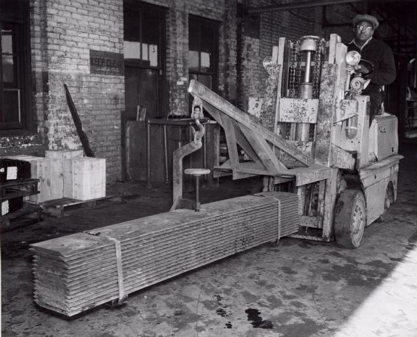 A factory worker at International Harvester's Tractor Works is using a forklift with a small crane to transport materials.