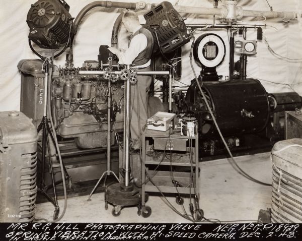 R.G. Hill taking photographs in the engine testing laboratory. The original caption reads: "R.G. Hill, chief photographer at Gas Power Engineering Department International Harvester Company, at Chicago, is taking pictures of valve spring vibration with a high-speed camera in the engine testing laboratory. The engine photographed by Mr. Hill is under dynamometer test for power and performance."