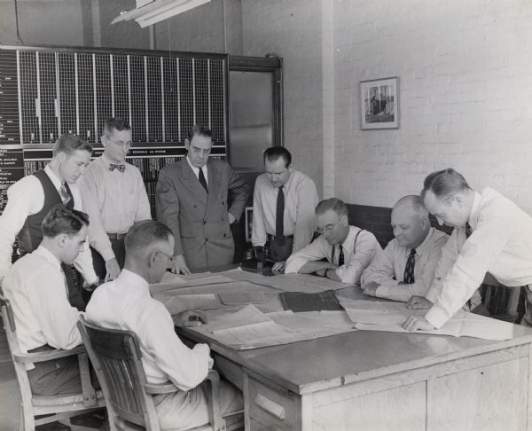 Heads of various divisions of International Harvester's truck(?) engineering department gather around a table to look over blueprints and plans. Clockwise, the men in the photograph are: J.C.A. Straub, engine design; D.J. Renno, body design; W.J. Fisher, service engineer; P.T. Brantingham, assistant chief engineer; H.K. Reinoehl, chief engineer; W.R. Westphal, experimental engineer; Tom Carney, new development; J.A. Bundy, transmission and axles; and S.G. Johnson, chassis.