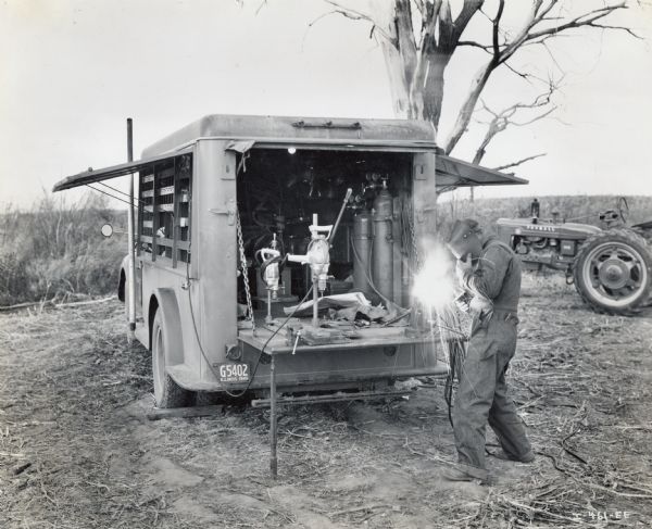 A man welding in the back of a mobile machine shop out in a field near a tractor is able to modify parts of experimental harvesting machines on location. A Farmall tractor is in the background. The original caption reads: "With this mobile machine shop International Harvester engineers and designers are able to modify experimental parts of harvesting machines or make new ones right in the field, as they follow the Harvester "caravans" northward from beginning to end of the harvesting seasons. The farms of North Carolina are the proving grounds for the Company's harvesting machines."