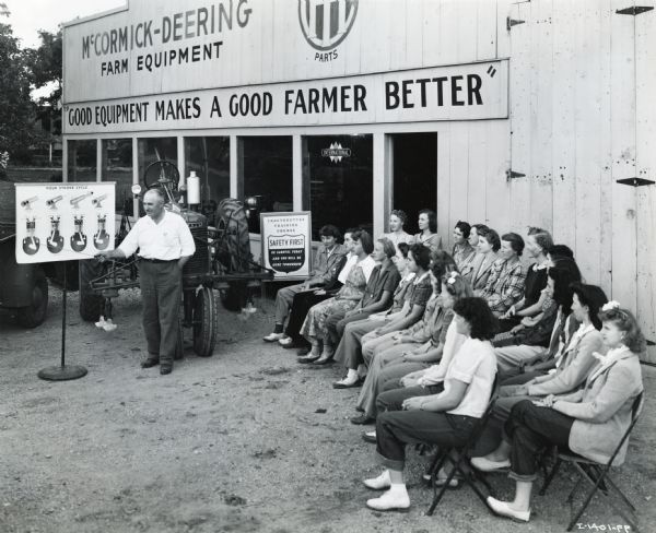 A group of seated young women at "Tractorette" School are instructed by John Schneider outside Nodaway County Implement Company, an International Harvester dealership.