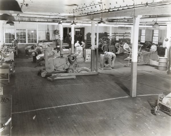 Men are standing among machines on the assembly floor in one of the International Harvester Company's engineering shops. The original caption reads: "Assembly floor in one of International Harvester Company's engineering shops in Chicago, where the parts of experimental and pre-production machines are put together. In the foreground is a sample model of a corn sheller. In the background are roughage mills, feed grinders, and other experimental implements."