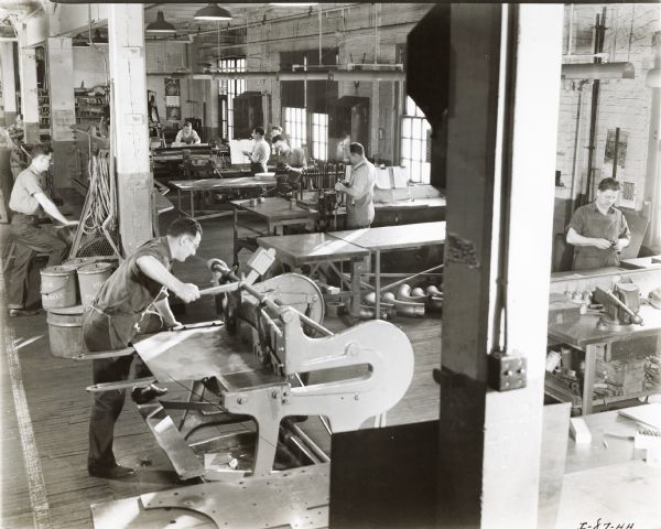 Men working in the sheet metal shop of the farm machinery engineering department of the International Harvester Company. The original caption reads: "Part of the sheet metal shop of the farm machinery engineering department of International Harvester Company at Chicago. The operator in the foreground is shearing sheet metal to dimensions called for by working drawing provided by the designer."