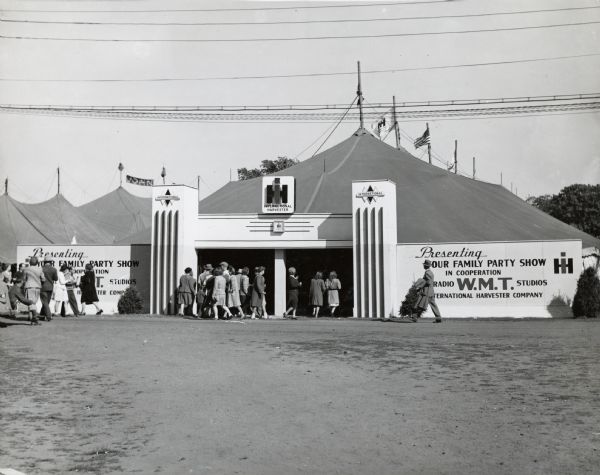 A group of people enter the International Harvester tent at the Dairy Cattle Congress. The sign on the outside of the tent reads: "Presenting Your Family Party Show in Cooperation with Radio W.M.T. Studios; International Harvester Company."