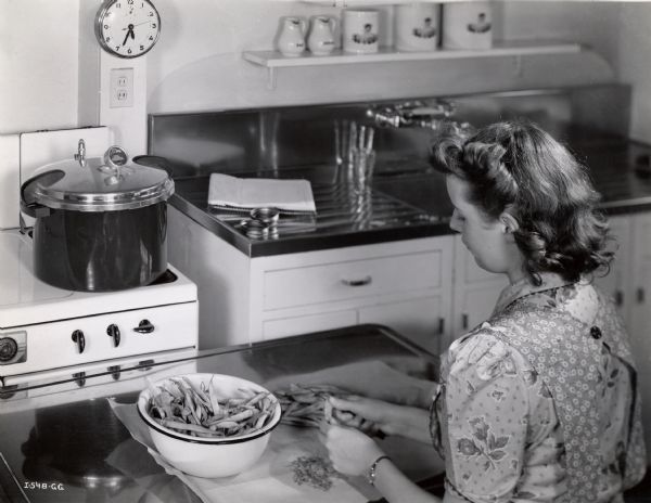 Elevated view of a woman cutting string beans at a kitchen counter. A pot on a stovetop and a sink are in the background.