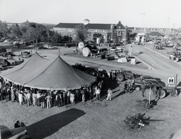 A crowd gathers underneath a tent near an International Harvester sign and farm machines at the Kansas State Fair. The sign on the building in the background reads: "4-H Club Exhibits" and the display behind the tent advertises Doran Tanks.