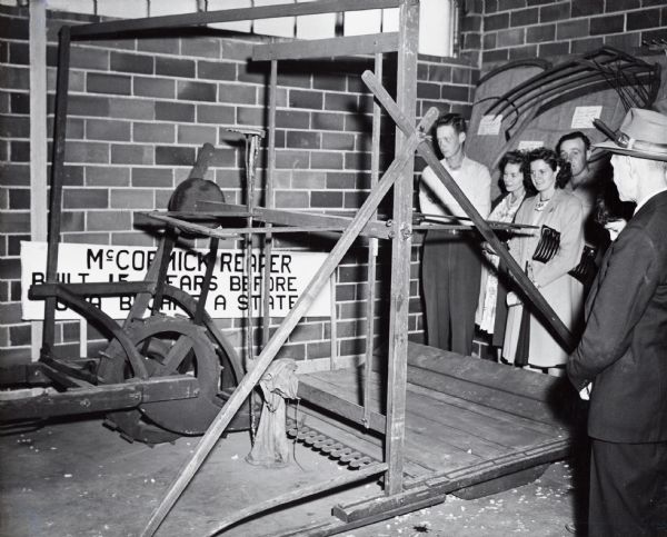 A group of onlookers gathers around a McCormick reaper replica display at the Iowa State Fair. The sign behind the reaper reads: "McCormick Reaper Built 15 Years Before Iowa Became A State."