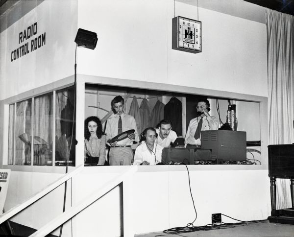 A group of radio broadcasters working from a radio control room at the Iowa State Fair. An International Harvester clock is mounted on the booth's outside wall.