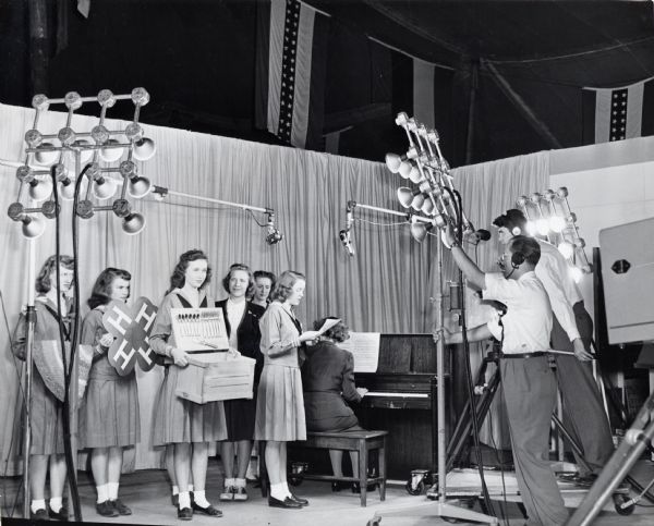 A group of 4-H members standing underneath lights to be filmed for television at the Iowa State Fair, while a woman nearby is playing the piano.