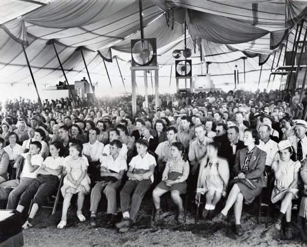 A group of spectators sits under a tent to watch a show at the Indiana State Fair. A sign in the background advertises "International Trucks."