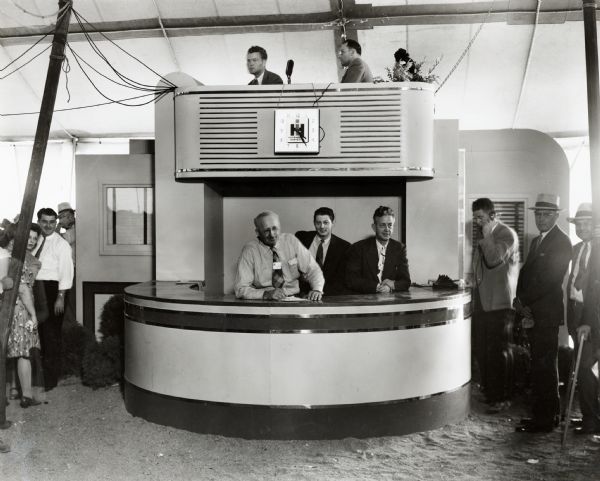 Men at the Indiana State Fair sit behind the lower counter of an International Harvester booth while others sit above with a microphone.