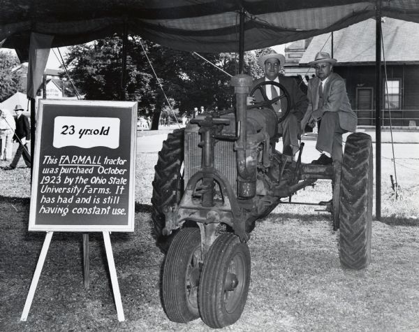 Two men pose on a twenty-three year old Farmall Regular tractor on display under a tent at the Ohio State Fair. The sign reads: "23 yrs. old; This Farmall tractor was purchaed <i>[sic]</i> October 1923 by the Ohio State University Farms. It has had and is still having constant use."