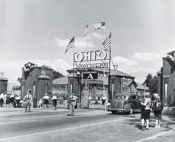 Police direct traffic through "Entrance A" at the Ohio State Fair while pedestrians walk nearby. Woman in uniforms sell concessions outside the entrance.