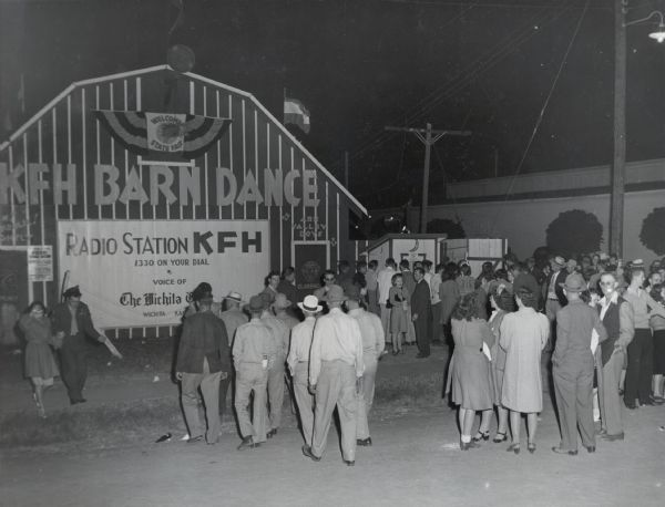 A crowd gathers outside a barn advertising the "KFH Barn Dance" at the Kansas State Fair. A sign on the barn reads: "Radio Station KFH; 1330 On Your Dial; Voice of The Wichita...(?); Wichita, Kansas."  The Wichita branch of International Harvester Company estimated that 50,000 of 250,000 total fairgoers visited their display.