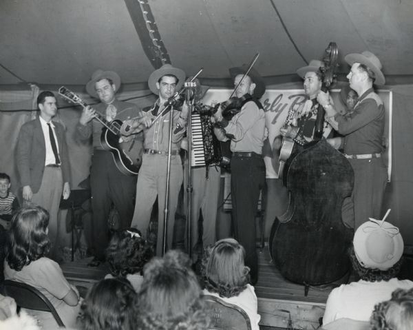A musical group comprised of men playing guitars, fiddles, an accordion, and a string bass perform on stage at the Louisiana State Fair.