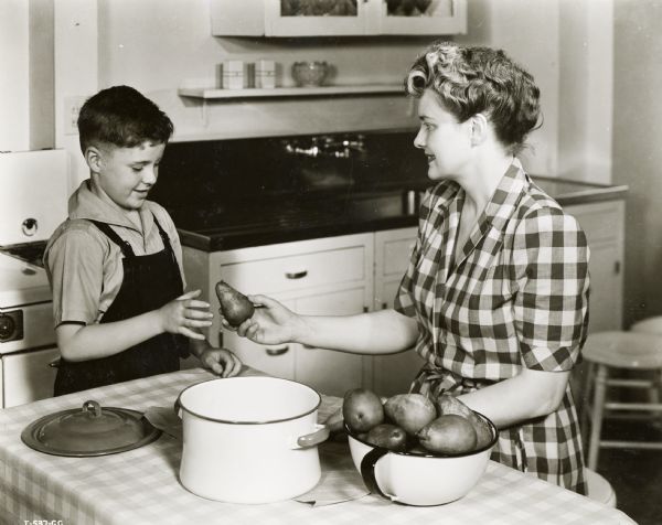 A woman sitting at a kitchen table is handing a pear to a young boy standing next to her.
