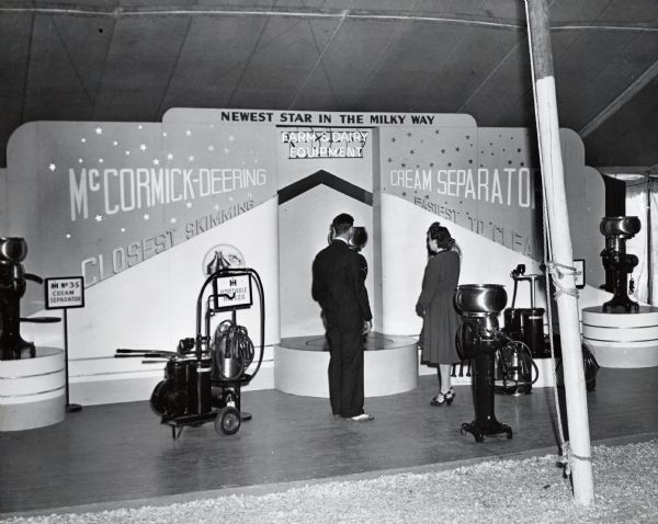 A couple looks at the McCormick-Deering display of cream separators and milking machines at the Iowa State Fair. The display text reads: "Newest Star in the Milky Way," "Farm & Dairy Equipment," "McCormick-Deering Cream Separator," and "Closest Skimming - Easiest to Clean."