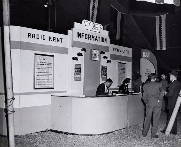 A man and a woman sit behind the International Harvester information booth at the Iowa State Fair. An International Harvester clock hangs on the booth's wall, as well as advertisements reading: "Radio KRNT" and "RCA Victor", and a poster reading: "Why We Are Not Displaying Our Full Line of Equipment."