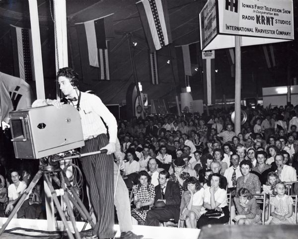 A television crew worker films "Iowa's First Television Show" while an audience looks on. The sign reads: "Iowa's First Television Show In Cooperation With Radio KRNT Studios, International Harvester Company."