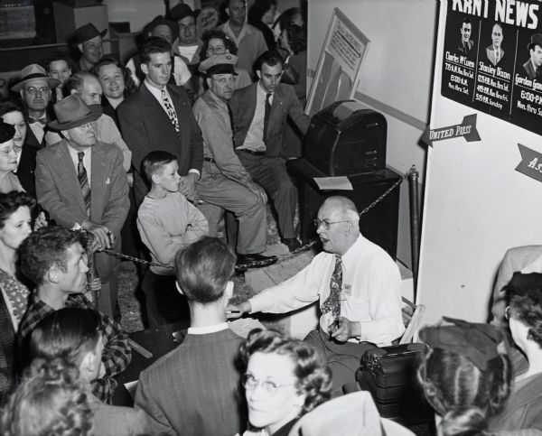 A crowd gathers around a KRNT news broadcaster at the Iowa State Fair. The sign behind the broadcaster reads: "Charles McCuen, Stanley Dixon, Gordon Gamm...(?)"