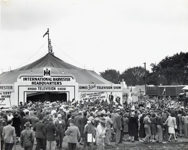 A large crowd gathers outside the International Harvester Headquarters tent to watch a broadcast of a radio television show. The signs on the outside of the tent read: "Iowa's First Television Show" and "See Popular Iowa Features, Fair Winners, KRNT Radio Stars Televised!"