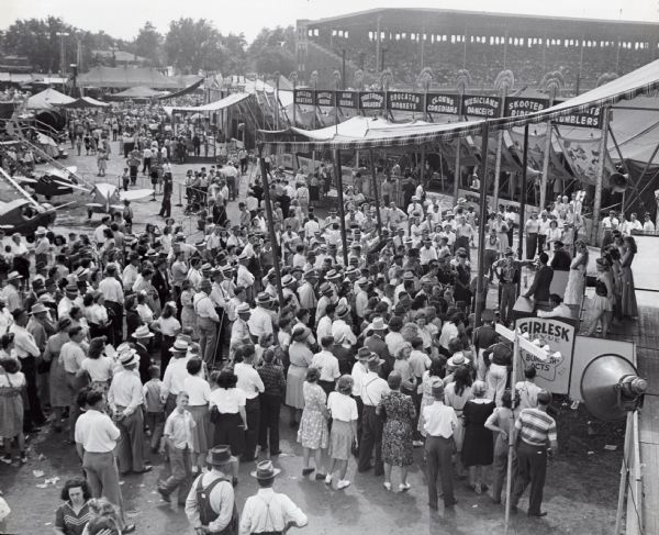 Elevated view of an audience gathering around the "Girlesk Revue" stage at the Iowa State Fair. The signs in the background read: "Roller Skaters; Bicycle Riders; High Divers; Tight Rope Walkers; Educated Monkeys; Clowns, Comedians; Musicians, Dancers; Skooter Riders;(?)...Tumblers."