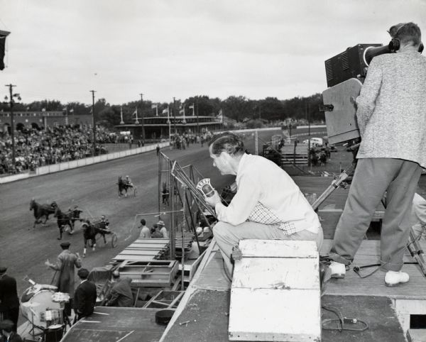 A KRNT radio broadcaster sitting on a platform above a horse track at the Iowa State Fair as another man on the right is filming the race for television. A band is sitting near the track at bottom left.