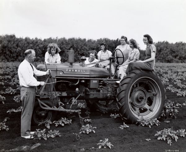 A group of female "Tractorette" class participants with their male instructor gather around a Farmall tractor. The original caption reads: "Part of tractorette class conducted by Day & Perkins of Batavia, New York, was photographed on big truck farm in a muckland section near Elba operated by Porter & Bonney."