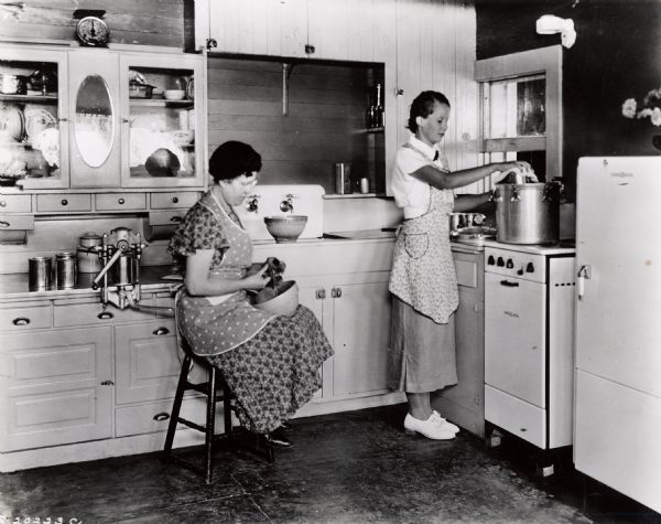A woman sitting on a stool in a kitchen peeling fruits or vegetables. Another woman is lifting the lid off a pot on the stove.