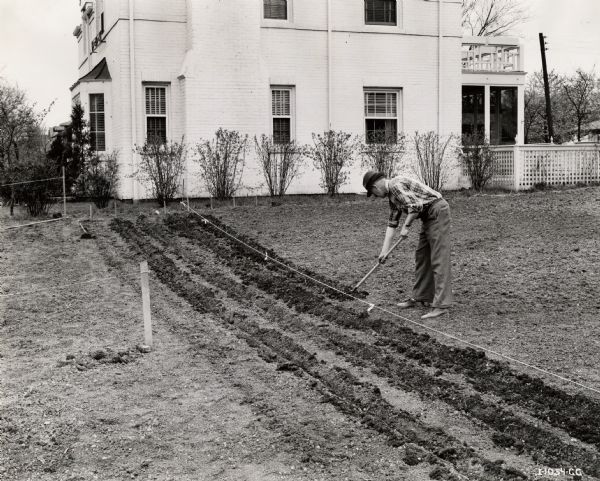 A man standing behind a house uses a hoe to loosen dirt for garden rows.