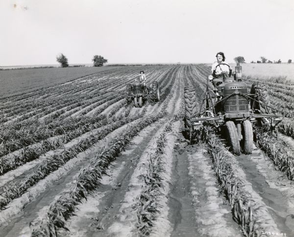 Two women on Farmall tractors with attached cultivators in a farm field.