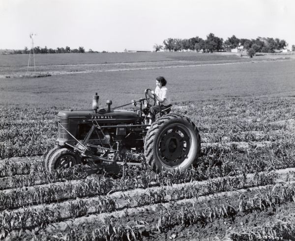 A woman operating a Farmall H tractor with an attached cultivator in a farm field.