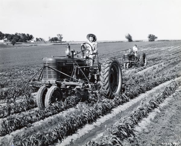 A woman is steering a Farmall H tractor through a farm field, while another person driving a tractor is following closely behind.