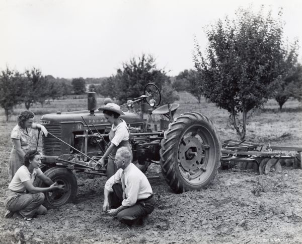 Ruth Sager stoops to speak to Wood Hendrickson, manager of C.O. Smith, an International Harvester dealership. Two women stand behind Sager and Hendrickson, examining a Farmall H tractor. The original caption reads: "On the very unusual 208-acre Hechalutz Training Farm near Hightstown, New Jersey, young Jewish women are training to be farmers in preparation for migration to hallowed Palestine."