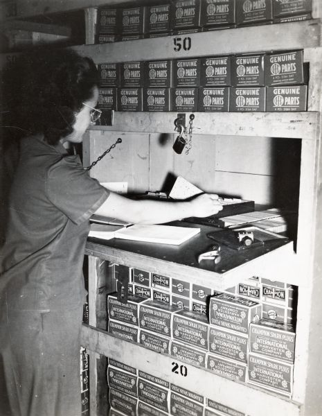 A female factory worker is looking at an index card in what appears to be a parts storage area at International Harvester's Tractor Works. The boxes stacked above the desk read "Genuine IHC Parts" and the boxes below read "Champion Spark Plugs."