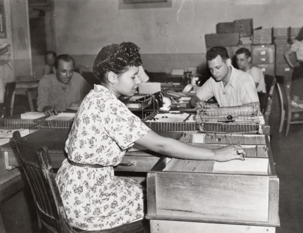 A woman at International Harvester's Tractor Works is looking through a filing cabinet or a card catalog. Other people are working in the background.