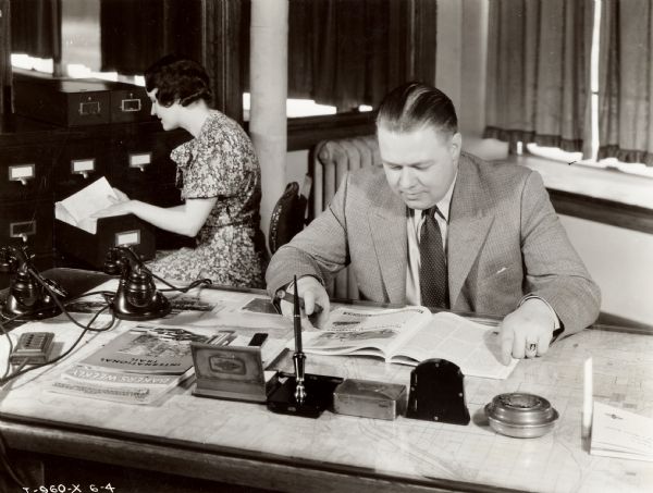 A man holding a cigar is sitting at a desk looking at an International Harvester trucks booklet. A copy of <i>International Trail</i> magazine is on the desk. A woman behind him is looking through a filing cabinet. The two are likely in the office of an International Harvester truck dealership.