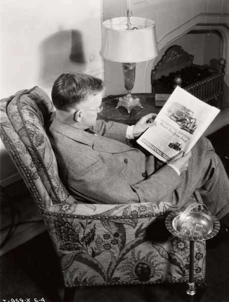 A man sits in an armchair reading an advertisement for the International Harvester "New 1/2-Ton Truck."  A free-standing ashtray is next to the chair.