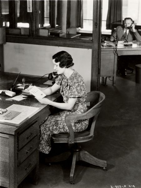 A woman at an office desk reads a pamphlet entitled, "What is Quality in a Motor Truck?" A male worker speaks on the telephone in the background. The two may be employed at an International Harvester dealership or branch house.