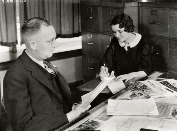 A man looks through papers at a desk while a woman, most likely a secretary, takes notes. The man and woman may have worked for an International Harvester dealership or branch house.
